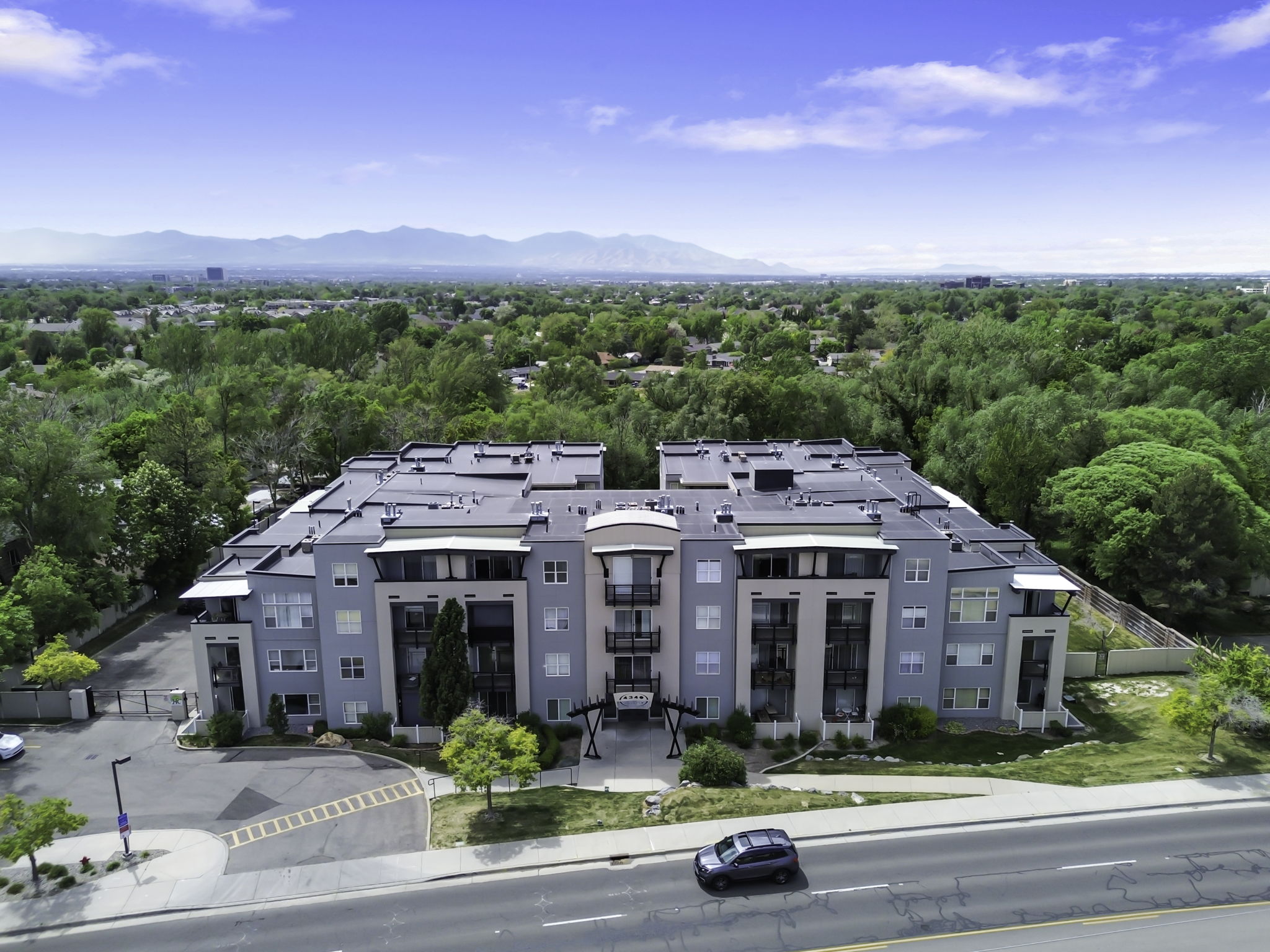 Modern multifamily building with a flat roofline, featuring a stone and stucco facade, private balconies, and a paved parking area