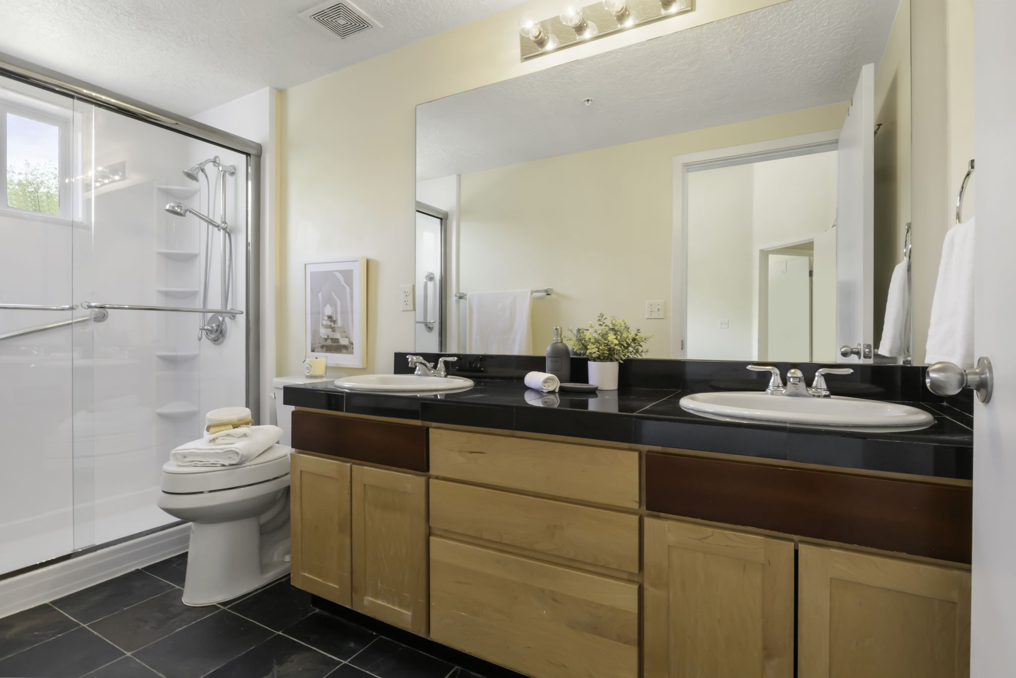 Bathroom featuring a dual vanity with a dark countertop and light wood cabinetry, a large mirror, a glass-enclosed shower with a built-in bench, a toilet, and dark tile flooring
