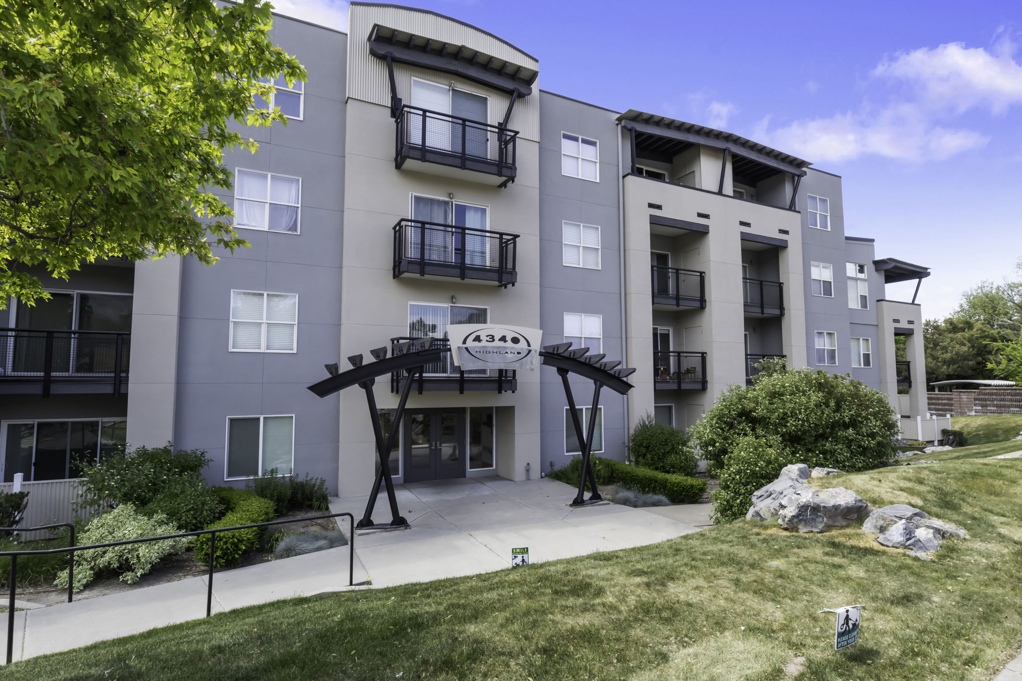 Contemporary multi-story building featuring a neutral color palette, multiple private balconies with black railings, a structured entryway canopy, a paved walkway, and surrounding landscaped greenery