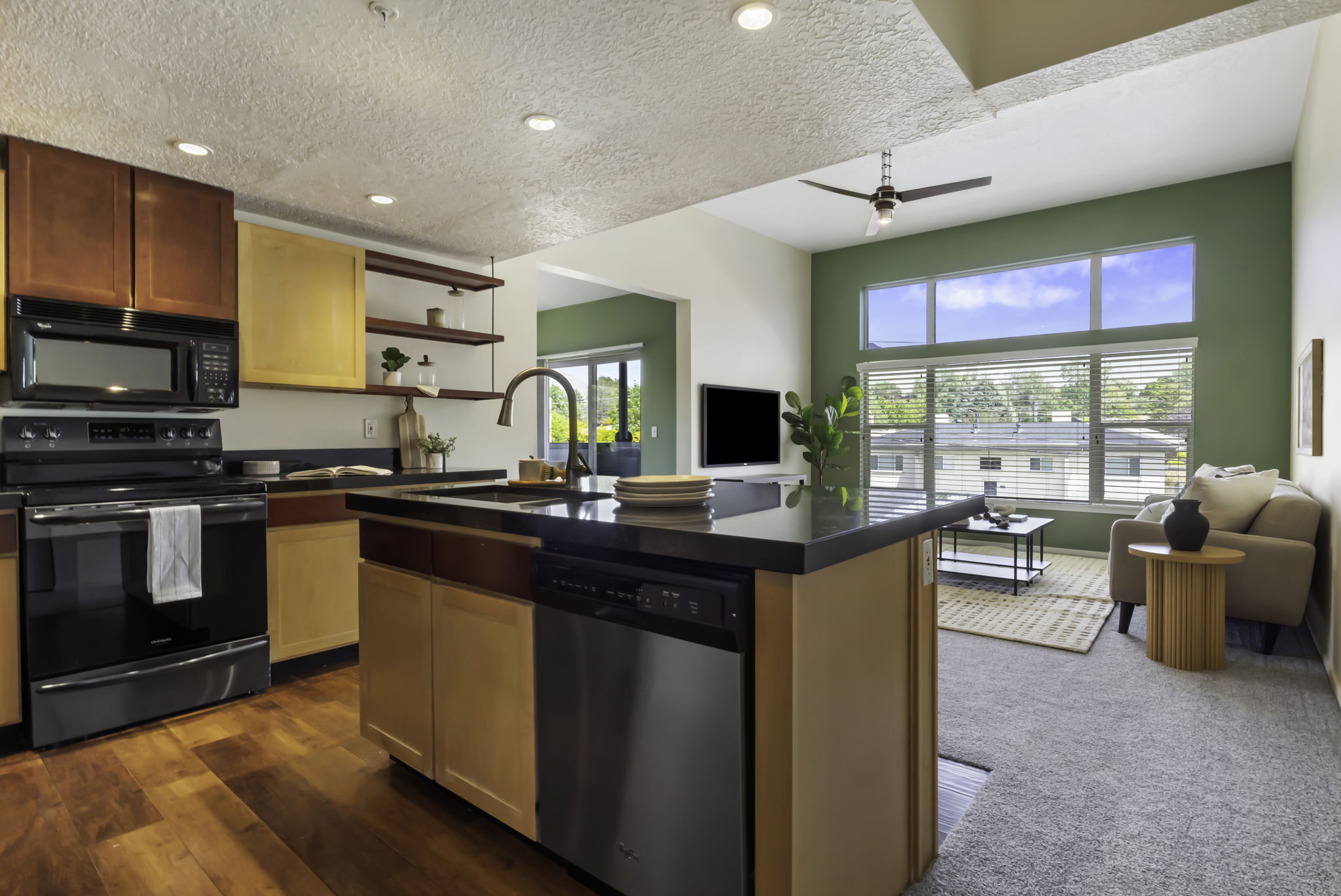 Open-concept kitchen and living area featuring wood-finish flooring in the kitchen, stainless steel appliances, a kitchen island with sink and integrated dishwasher, and a living area with large windows and a ceiling fan