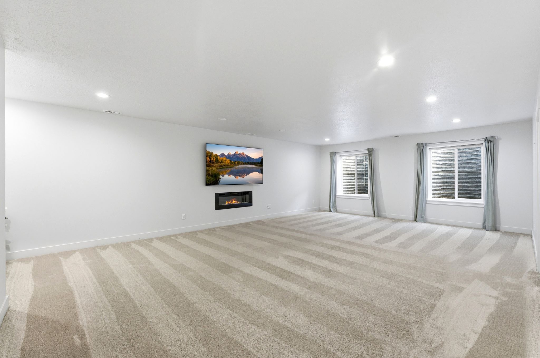 Spacious room featuring light-toned carpeting with subtle stripes, recessed lighting, and a wall-mounted linear fireplace