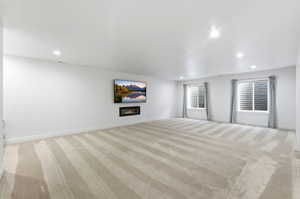 Spacious room featuring light-toned carpeting with subtle stripes, recessed lighting, and a wall-mounted linear fireplace