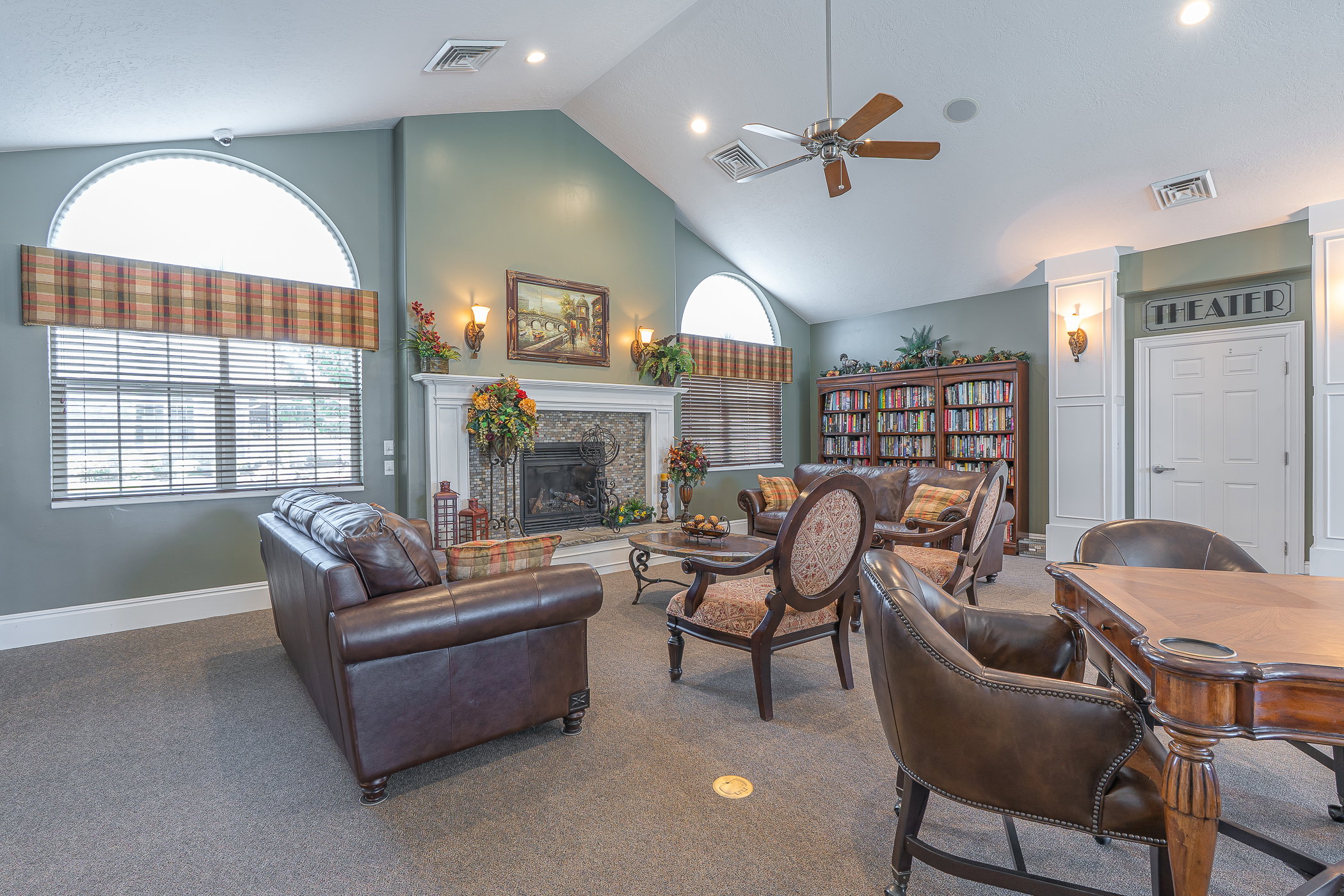 Spacious lounge area featuring a vaulted ceiling with a ceiling fan, a fireplace with a stone surround and white mantel, and multiple arched windows