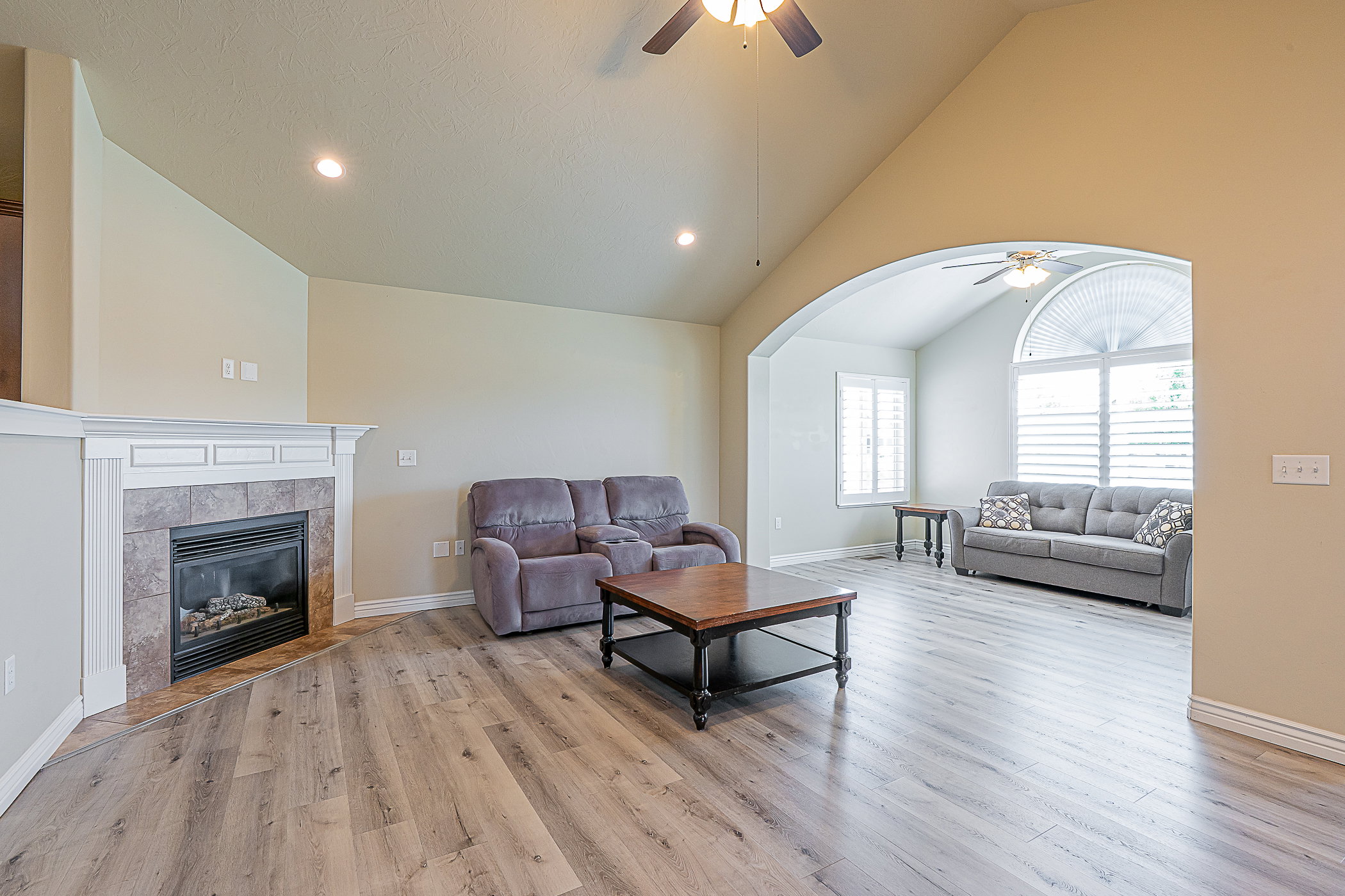 Spacious living area featuring wood-finish flooring, a corner fireplace with tile surround, vaulted ceilings, and an arched entry to an additional room with a half-round window and plantation shutters
