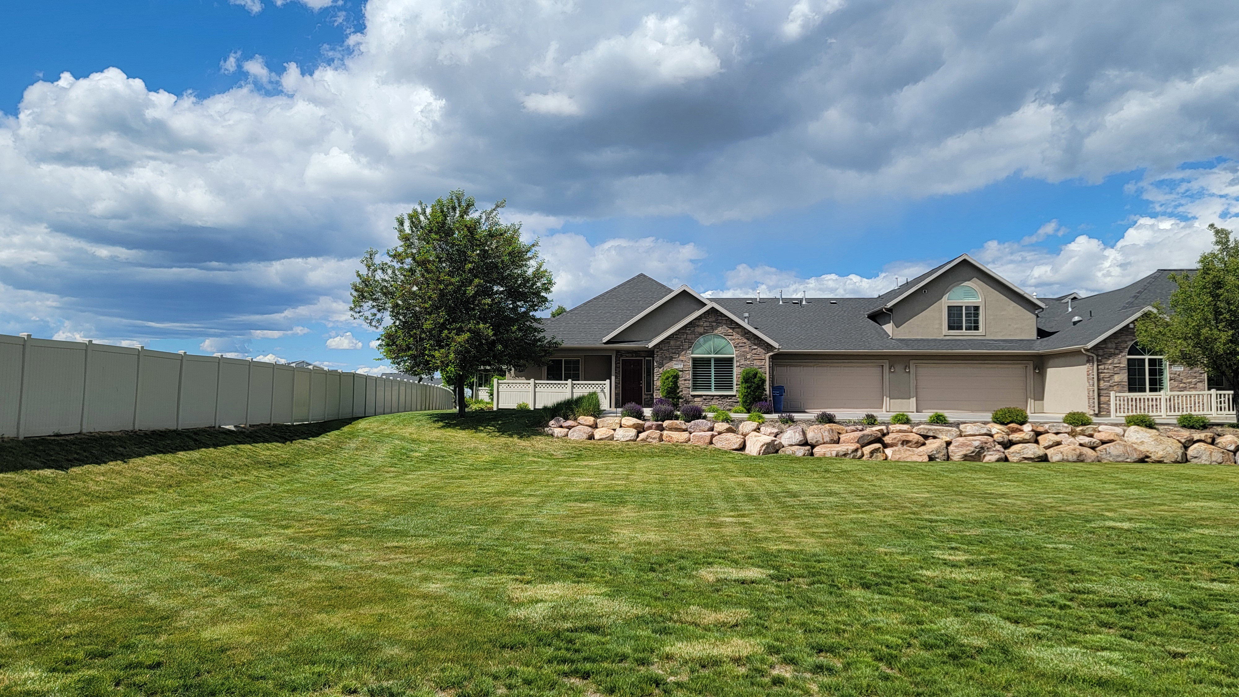 Spacious exterior featuring a large green lawn, stone and stucco facade, multiple garage doors, and a decorative rock retaining wall