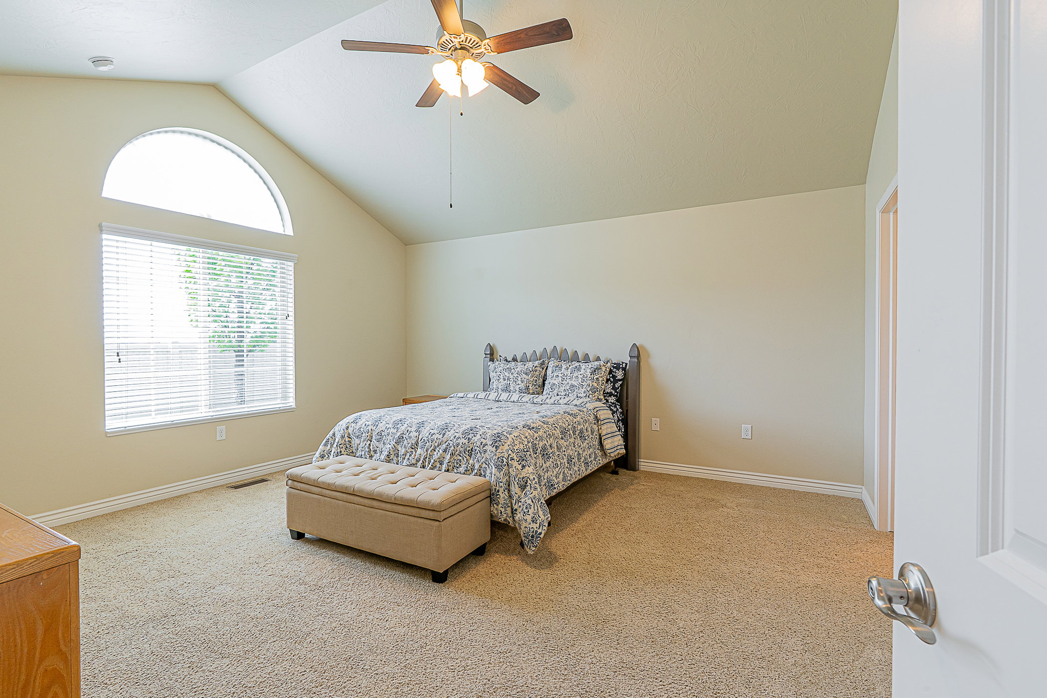 Vaulted ceiling room featuring a large arched window, a secondary window with blinds, a ceiling fan with integrated lighting, and neutral-toned carpeting