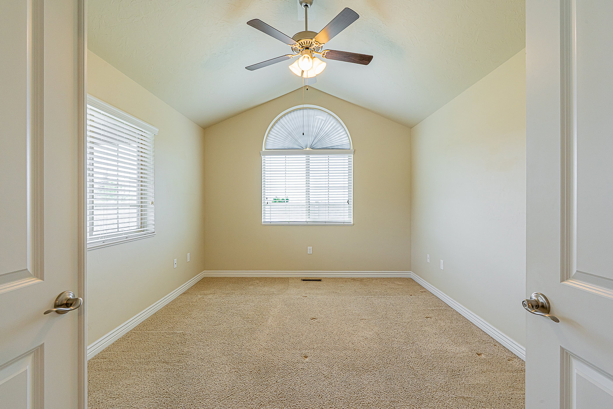 Vaulted ceiling room featuring a large arched window, an additional rectangular window with blinds, a ceiling fan with integrated lighting, and neutral tone carpet