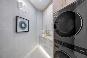 Integrated laundry area featuring a stacked washer and dryer, under-cabinet lighting, contemporary wall sconce, upper and lower cabinetry, and a light-toned countertop