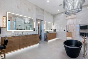 Spacious bathroom featuring a freestanding black soaking tub, dual wood-finish vanity cabinets with stone countertops, back-lit mirrors, and large format tile flooring