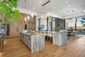 Open-concept kitchen and dining area featuring wood-finish flooring, two large stone islands, custom light wood cabinetry, and a full-height wine storage display