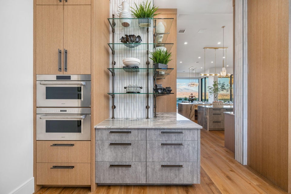 Built-in stainless steel double ovens, custom wood-finish cabinetry, gray textured lower cabinetry with dark hardware, glass shelving with dark metal supports, and a light-toned countertop