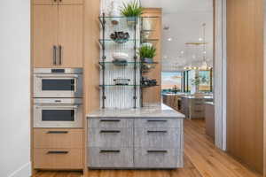 Built-in stainless steel double ovens, custom wood-finish cabinetry, gray textured lower cabinetry with dark hardware, glass shelving with dark metal supports, and a light-toned countertop