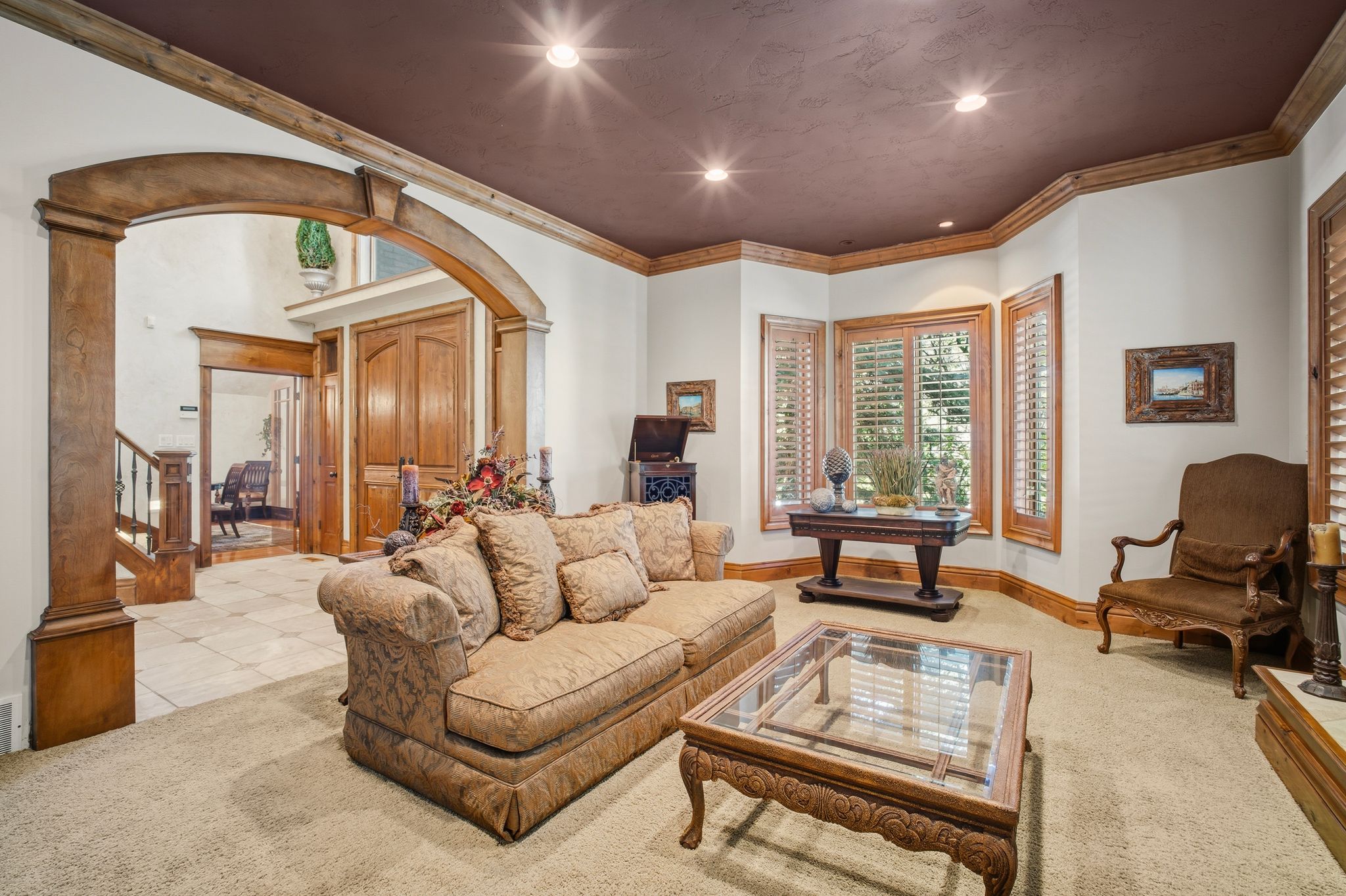 Living area featuring a tray ceiling with recessed lighting, bay windows with plantation shutters, wood trim accents, and an arched entryway leading to a tiled foyer with a double-door entry