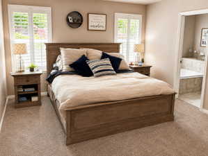 Carpeted room featuring dual windows with white plantation shutters, a wood-finish bed frame, and a tiled bathroom with a built-in tub visible through the doorway