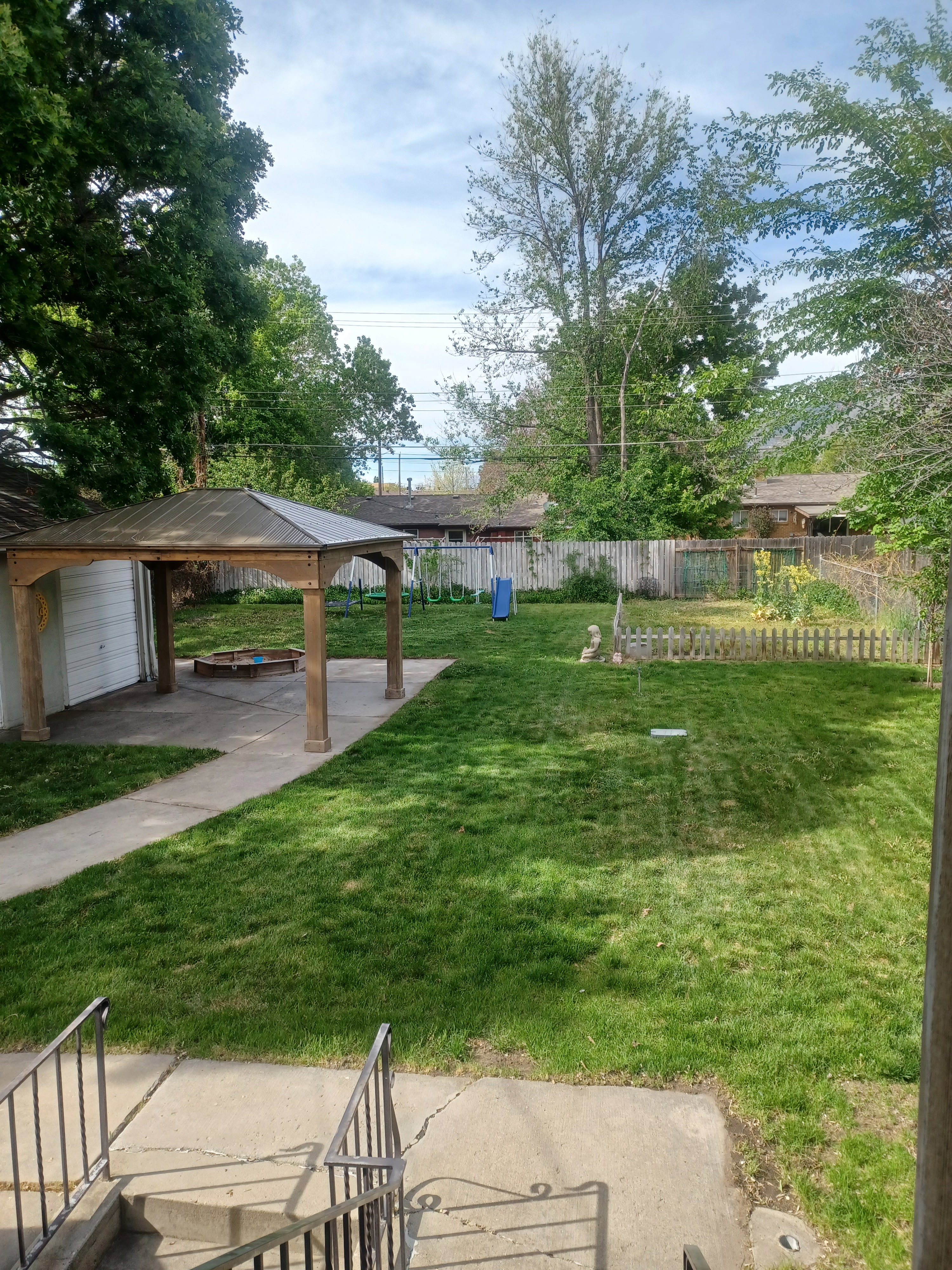 Spacious backyard featuring a manicured lawn, an open-sided gazebo with a metal roof, mature perimeter trees, and concrete pathways