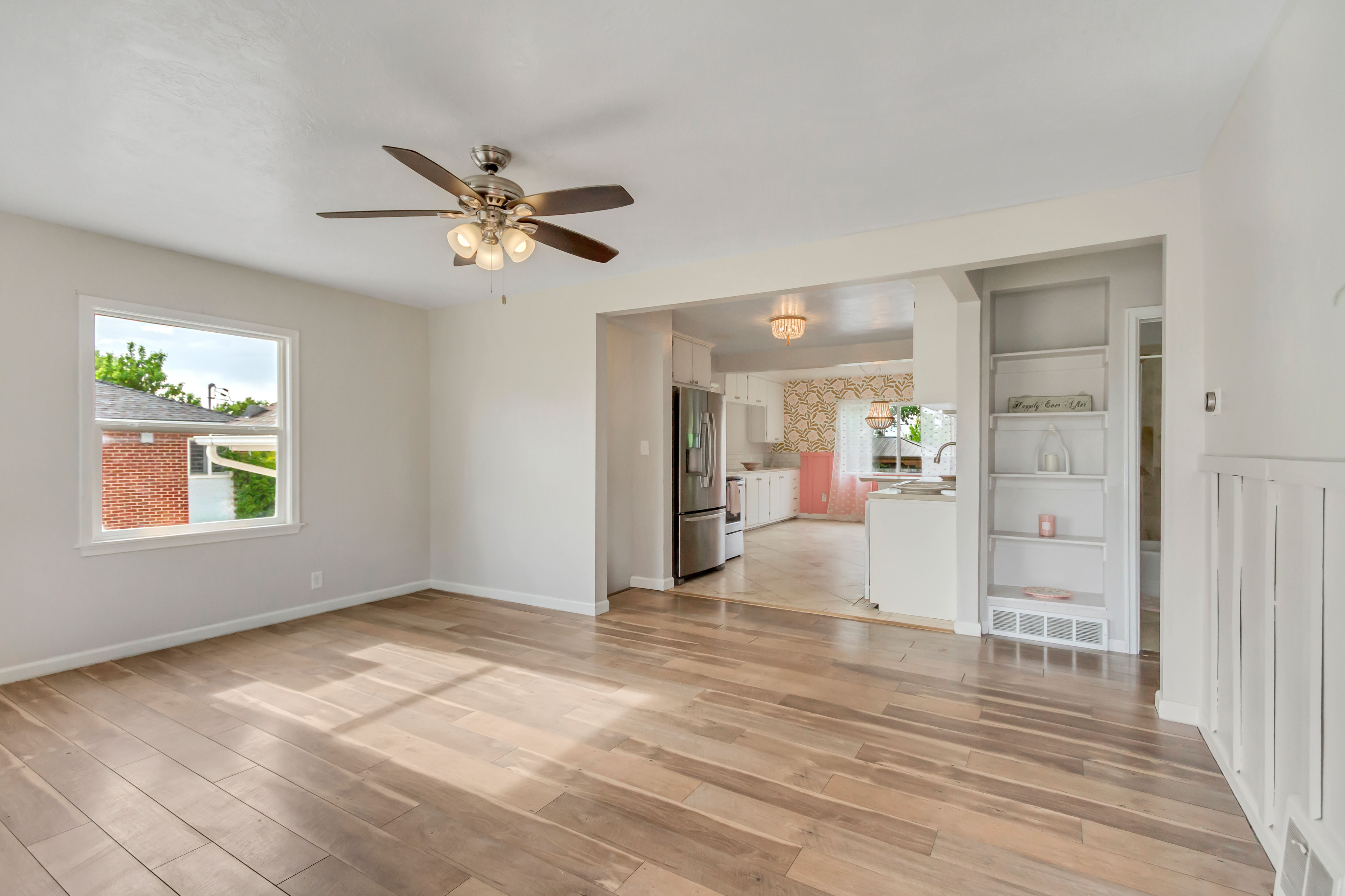 Spacious room featuring wood-finish flooring, a ceiling fan with integrated lighting, a bright window with white trim, built-in shelving, and white wainscoting