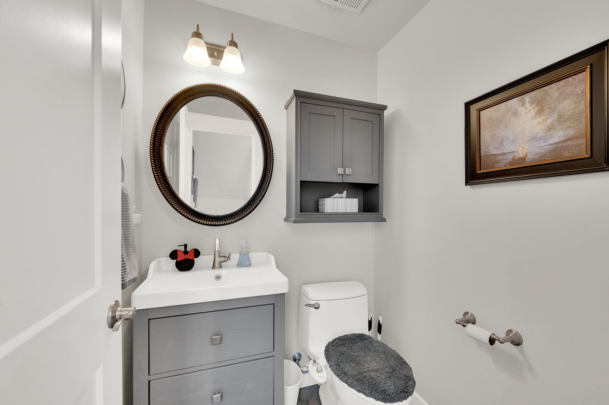 Powder room featuring a gray vanity with an integrated sink, a circular framed mirror, and a wall-mounted medicine cabinet with open shelving