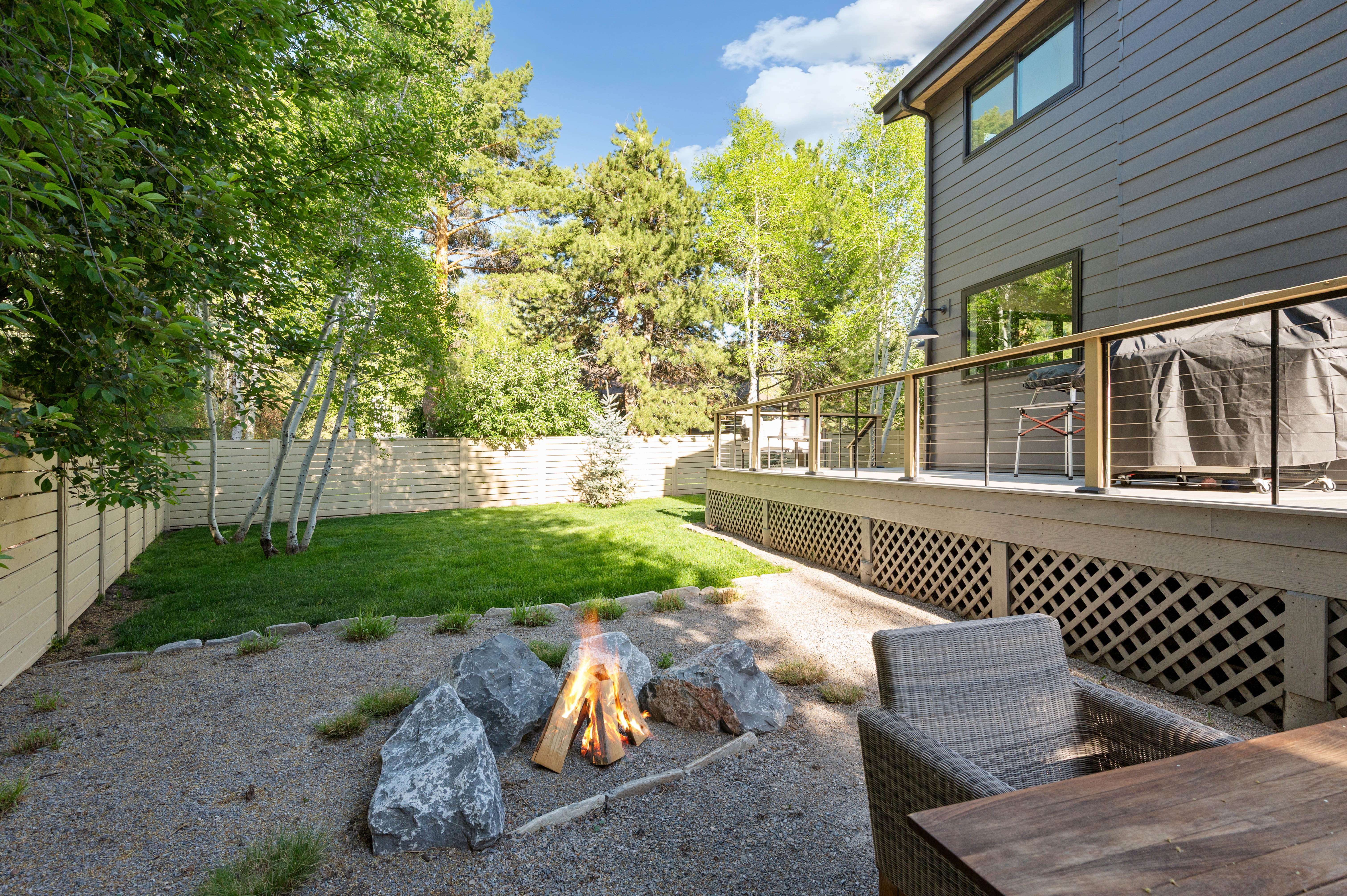 Outdoor area featuring a spacious deck with cable railing, a gravel patio with a rock fire pit, and a grassy lawn bordered by a privacy fence and mature trees