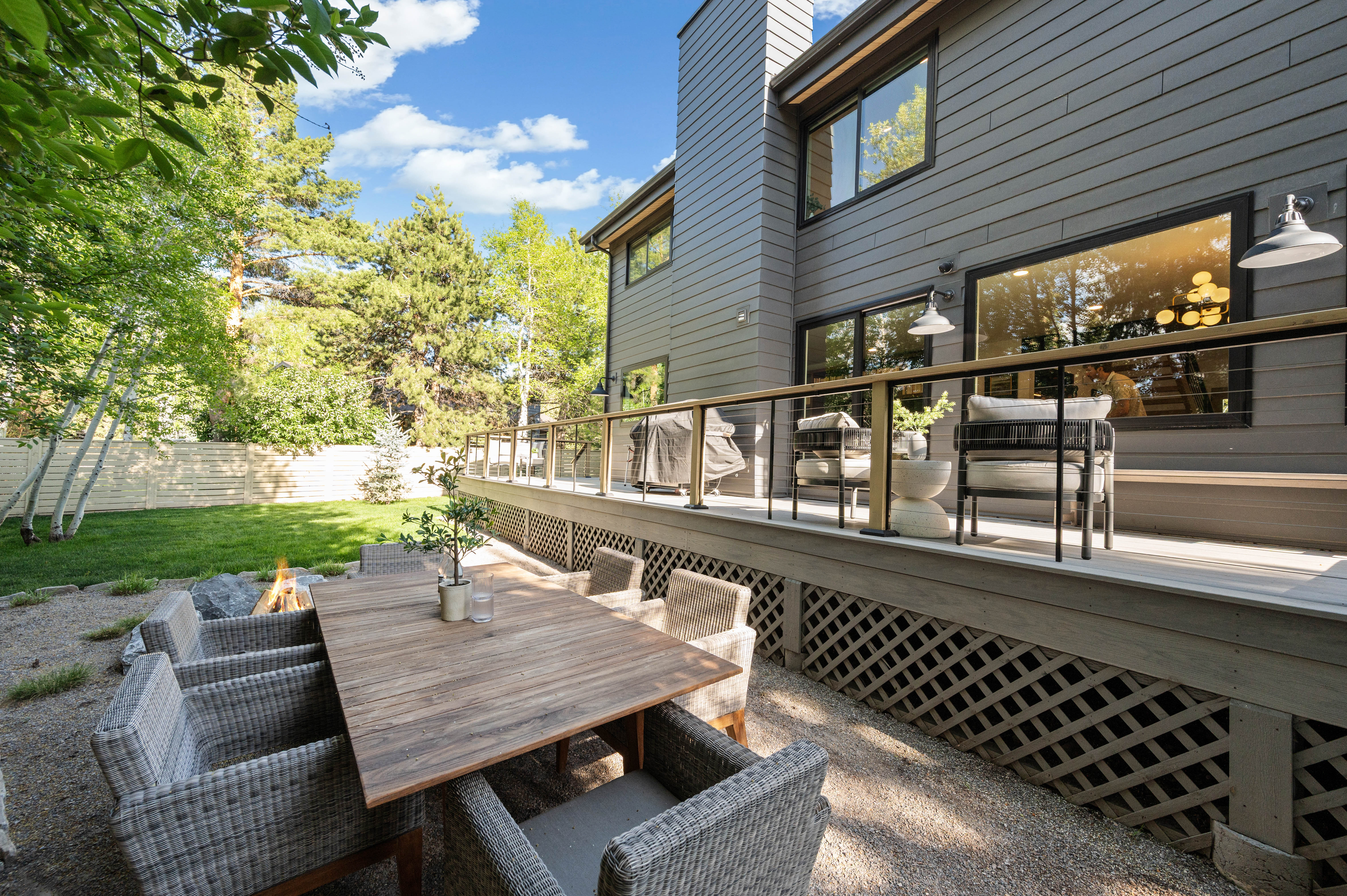 Expansive rear exterior featuring a multi-level deck with cable railing, a gravel patio with a built-in fire pit, mature trees, and a manicured lawn area