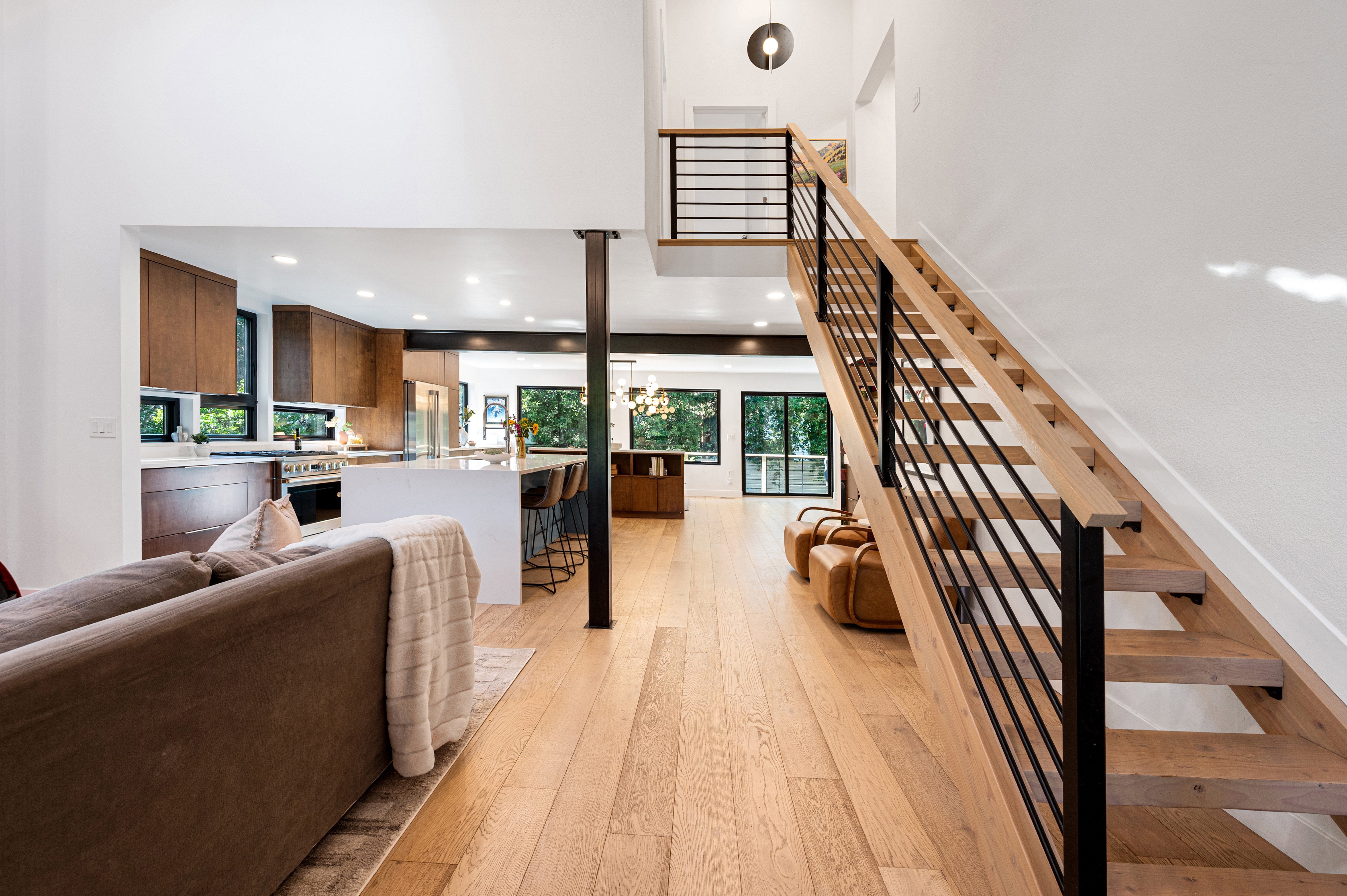 Open-concept interior featuring light wood-finish flooring, a modern staircase with wood treads and black metal railings, a kitchen with dark wood cabinetry and a white island, and a high ceiling with recessed lighting