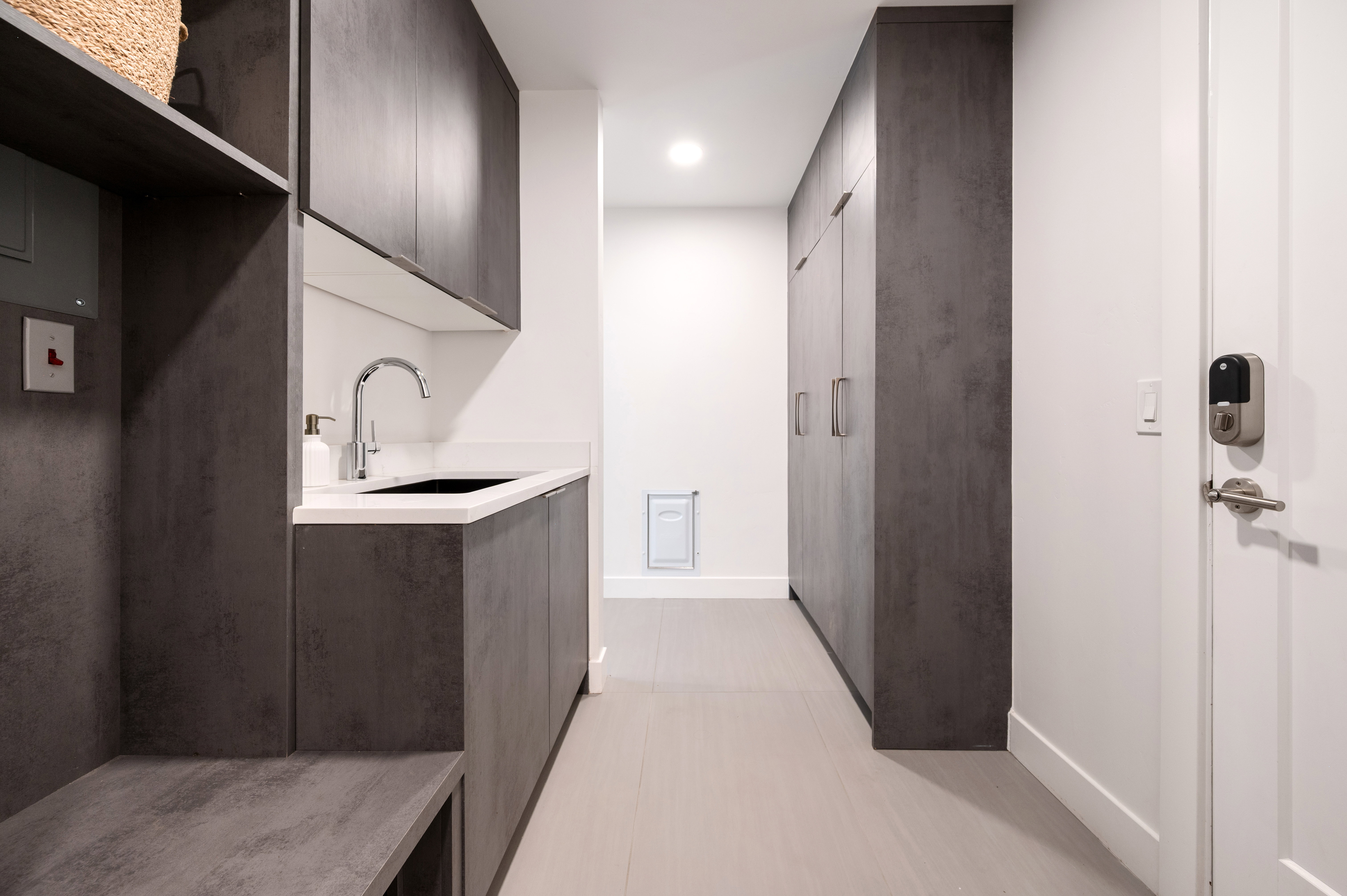 Streamlined utility space featuring extensive dark-finish cabinetry, integrated sink with chrome faucet, light wood-finish flooring, and white walls