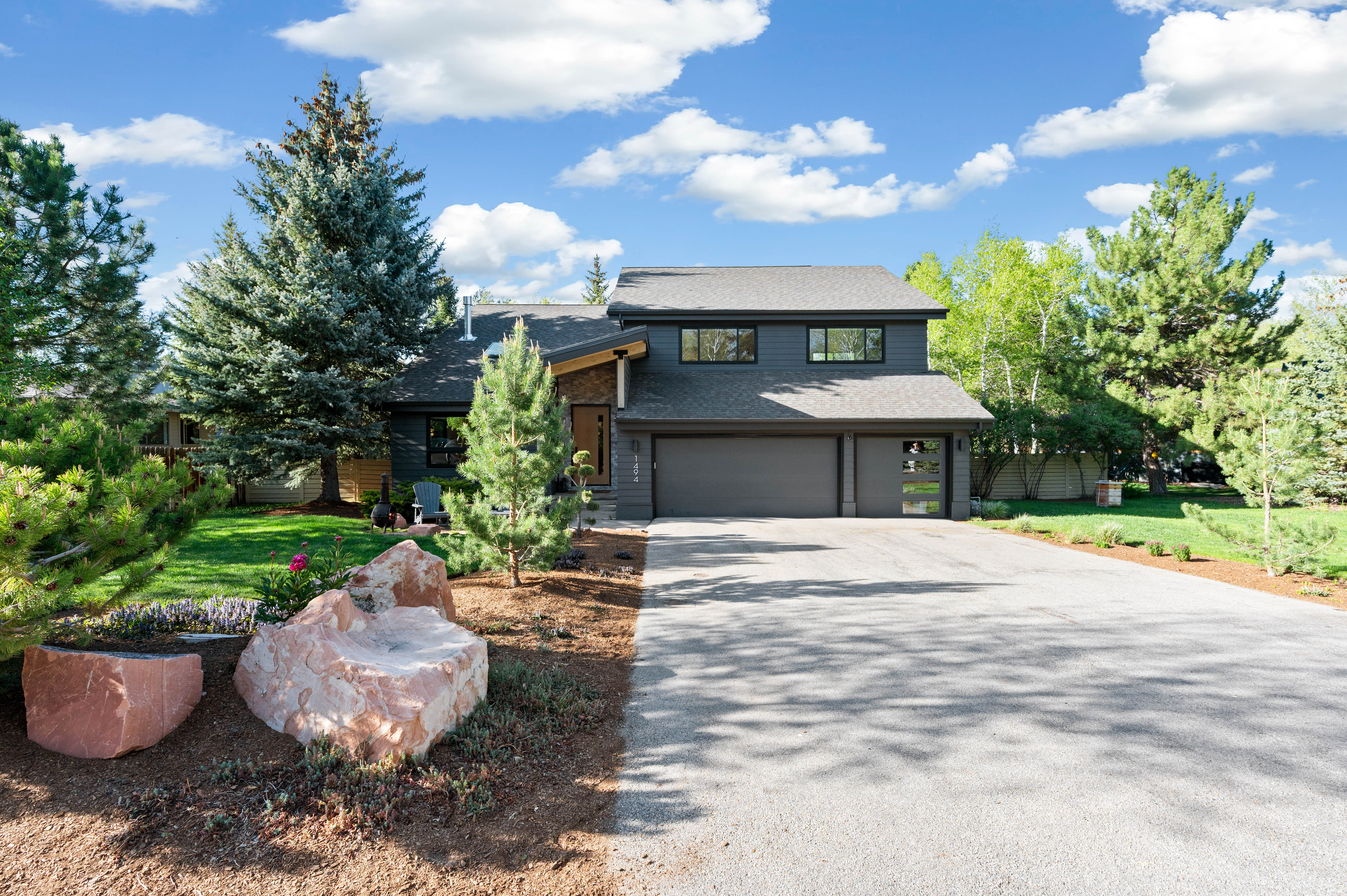 Contemporary gray exterior featuring a two-car garage, mature evergreen landscaping, and a paved driveway