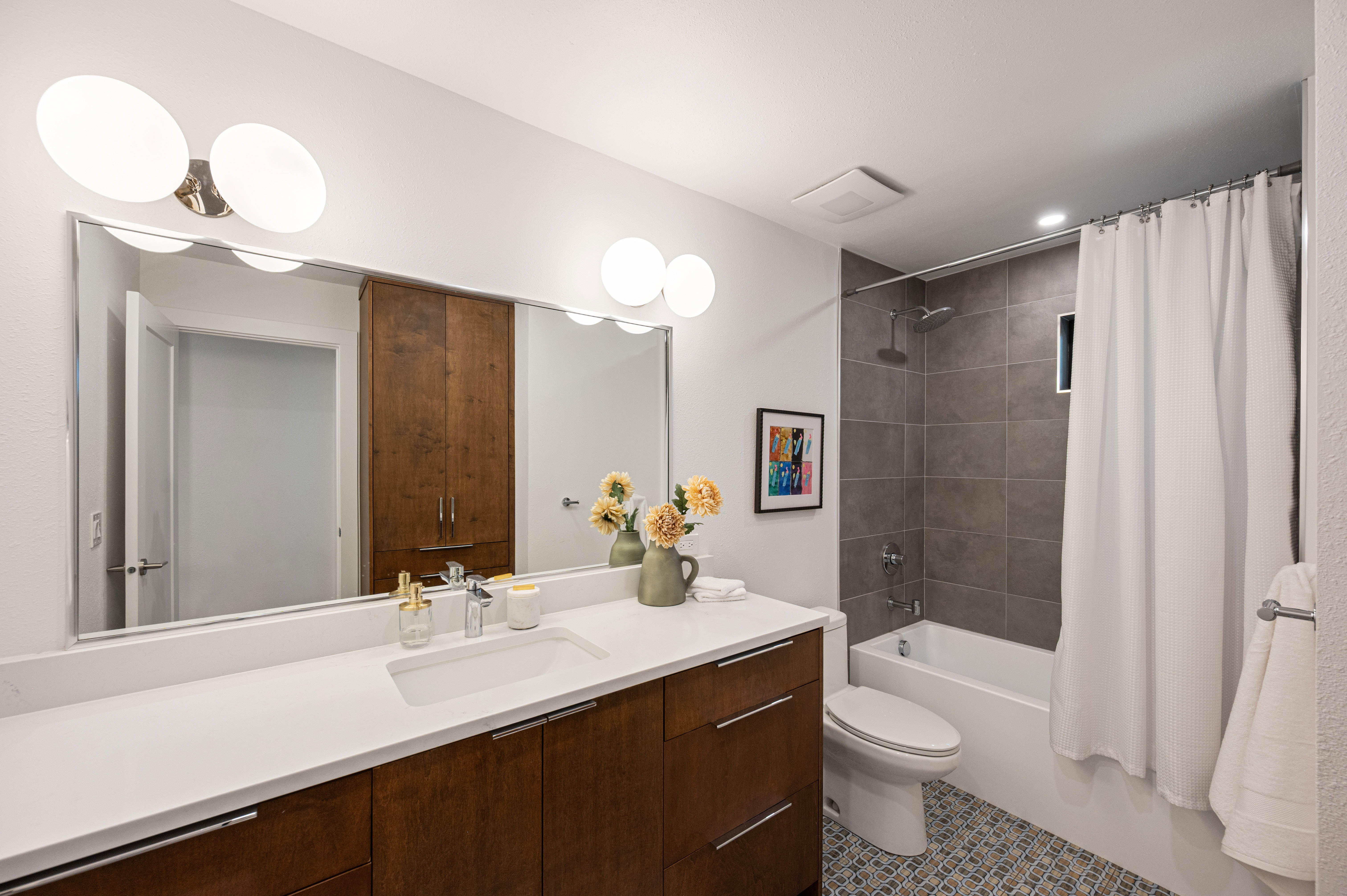 Bathroom featuring a wood-finish vanity with a white countertop and undermount sink, a large wall-mounted mirror, and a shower-over-tub with gray tile surround