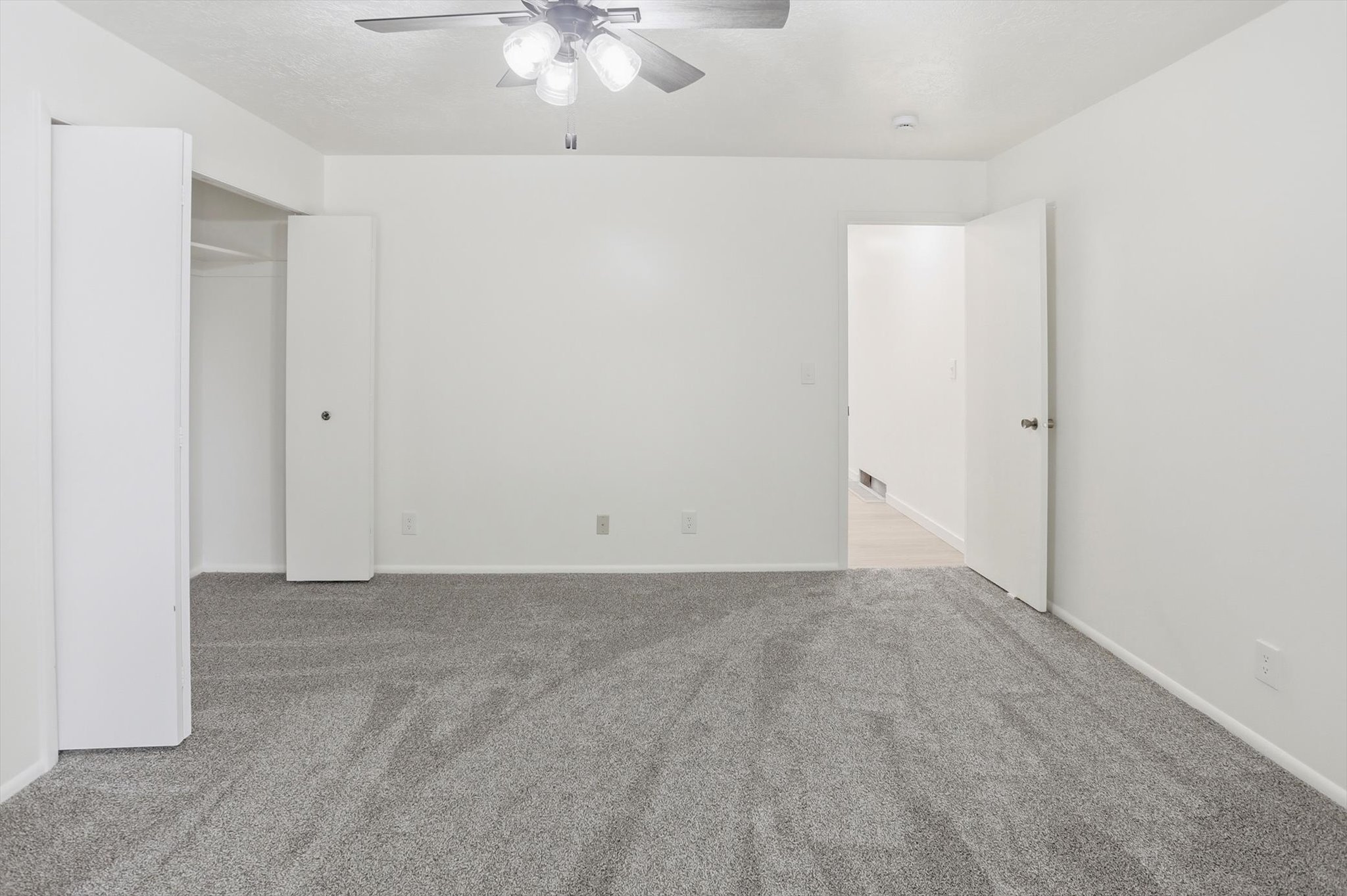 Spacious room featuring light gray carpeting, neutral white walls, and a ceiling fan with integrated lighting