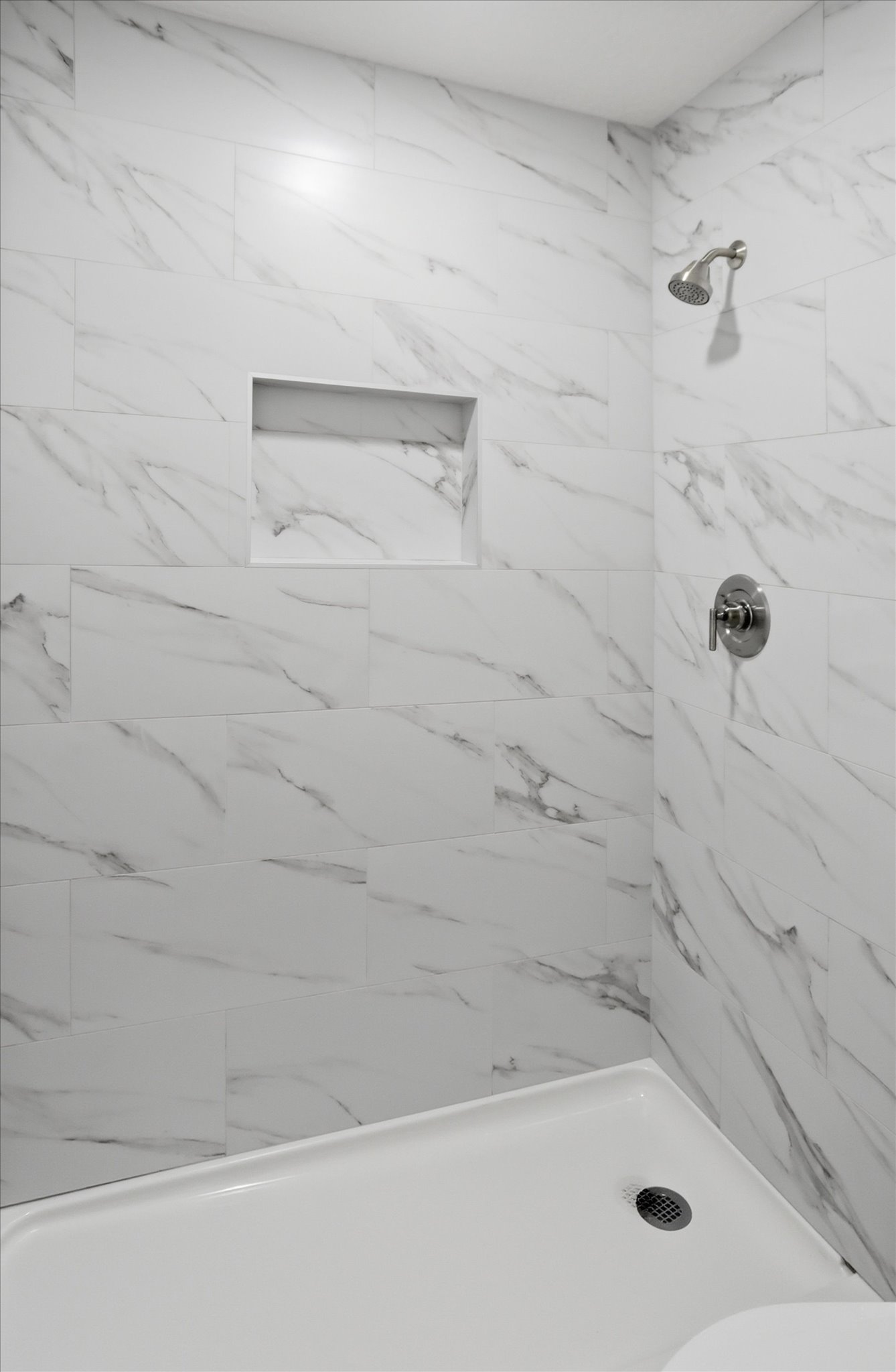 Shower featuring large format white tiled walls with gray veining, a built-in shower niche, and a brushed nickel shower head and control