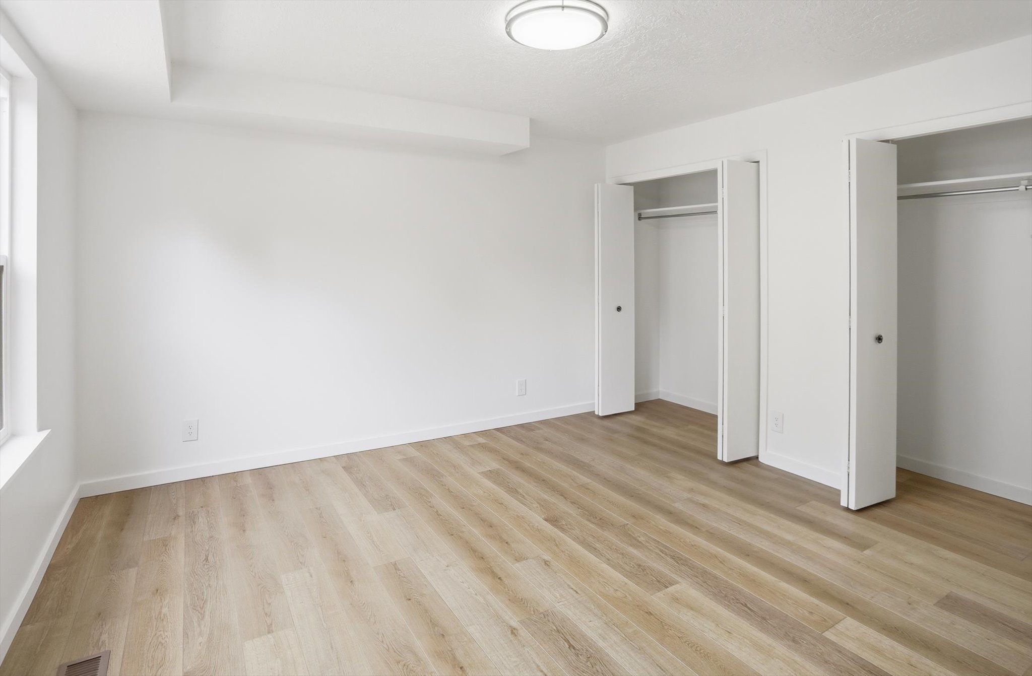 Spacious room featuring light wood-finish flooring, bright white walls, and a modern flush-mount ceiling light