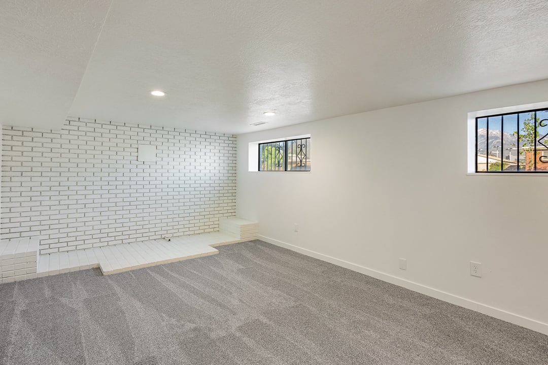 Spacious room featuring white painted brick wall with integrated hearth, neutral carpeting, recessed lighting, and two windows with decorative security bars