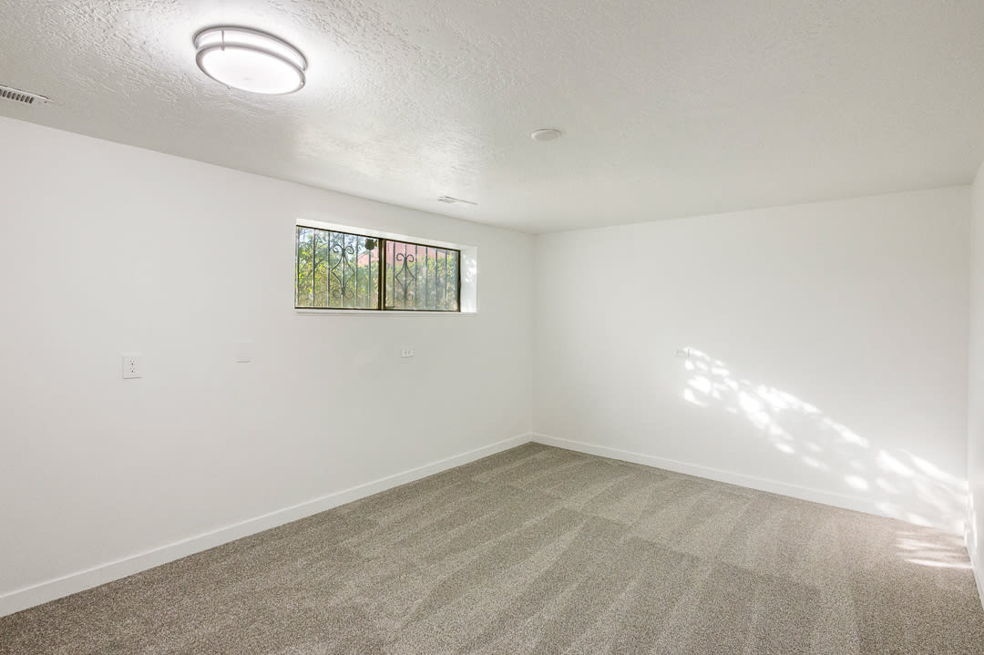 Carpeted room featuring white walls, a single window with decorative security bars, a flush-mount ceiling light fixture, and white baseboards