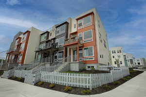 Modern townhome exteriors featuring varied siding materials and colors, contemporary window designs, multiple private balconies, individual front entrances with concrete steps, and white picket fences enclosing landscaped front yards