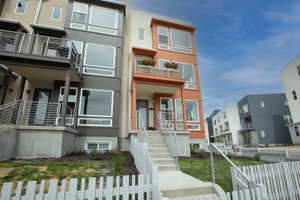 Modern townhome exterior featuring an orange facade, multiple balconies with metal railings, large windows, and a white picket fence