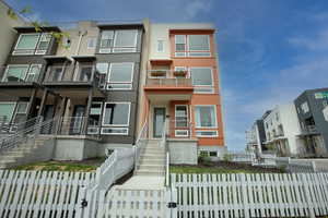 Modern townhome exterior featuring an orange accent facade, multiple balconies with metal railings, a white picket fence, and concrete entry stairs