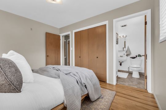 Bedroom featuring wood-finish flooring, two wood-paneled closet doors, and an open doorway revealing a bathroom with a pedestal sink and toilet