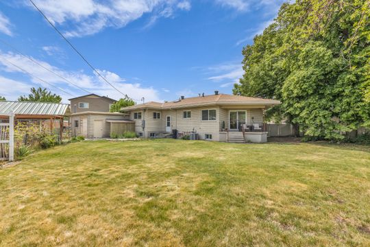 Rear exterior featuring a spacious lawn, covered patio, attached single-car garage, and mature perimeter trees