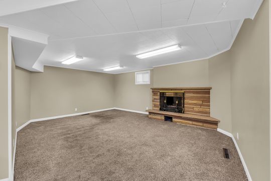 Spacious room featuring a corner fireplace with a stone surround and hearth, neutral-toned carpeting, and a drop ceiling with integrated fluorescent lighting