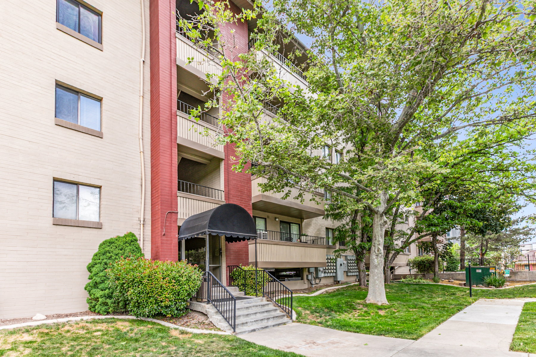 Multi-story building exterior featuring brick facade, private balconies, a covered entry with black awning, concrete steps with wrought iron railing, and established landscaping with mature trees