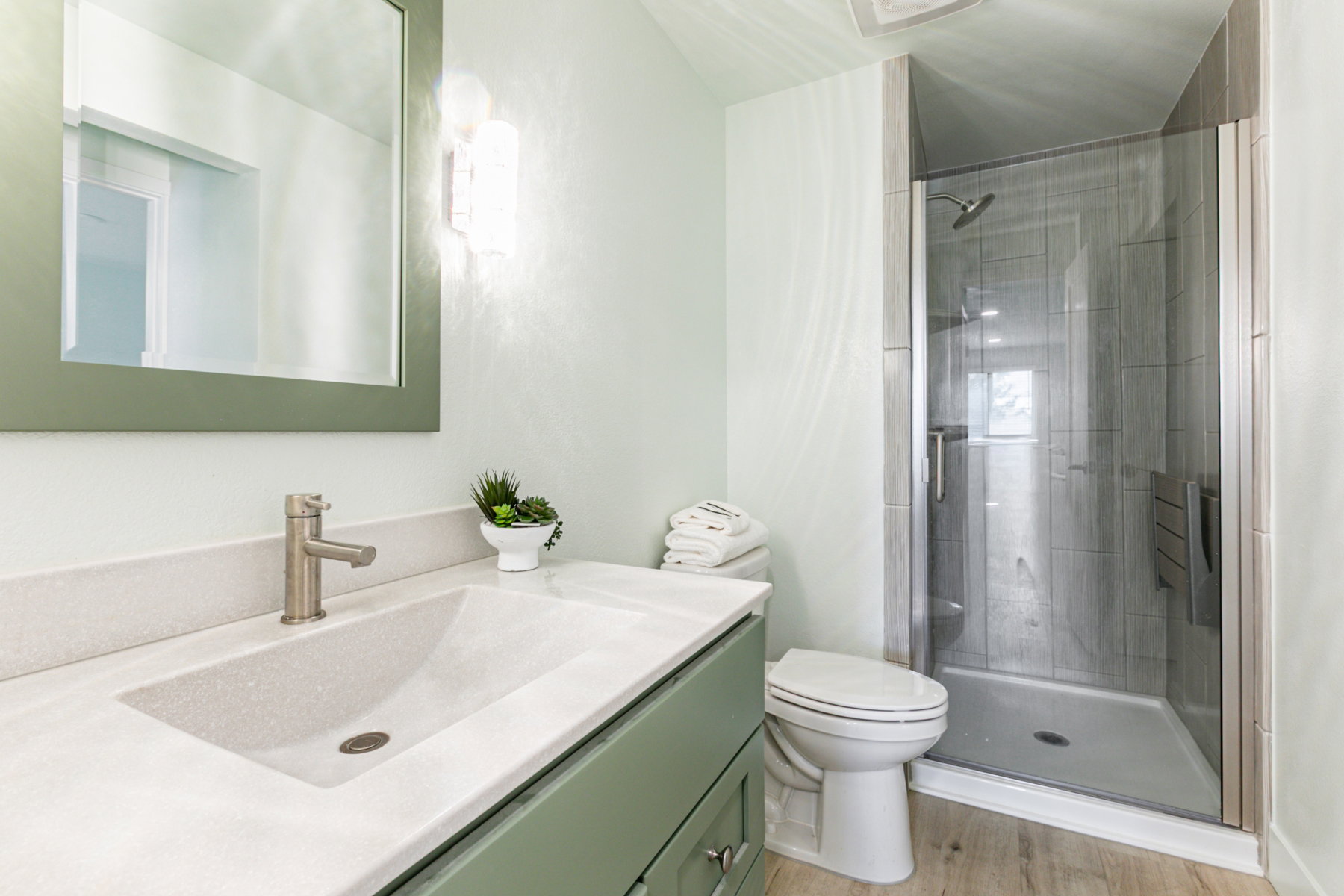 Bathroom featuring a glass-enclosed shower with tiled walls, a vanity with an integrated sink and light-colored countertop, green cabinetry, and a framed mirror with wall-mounted lighting