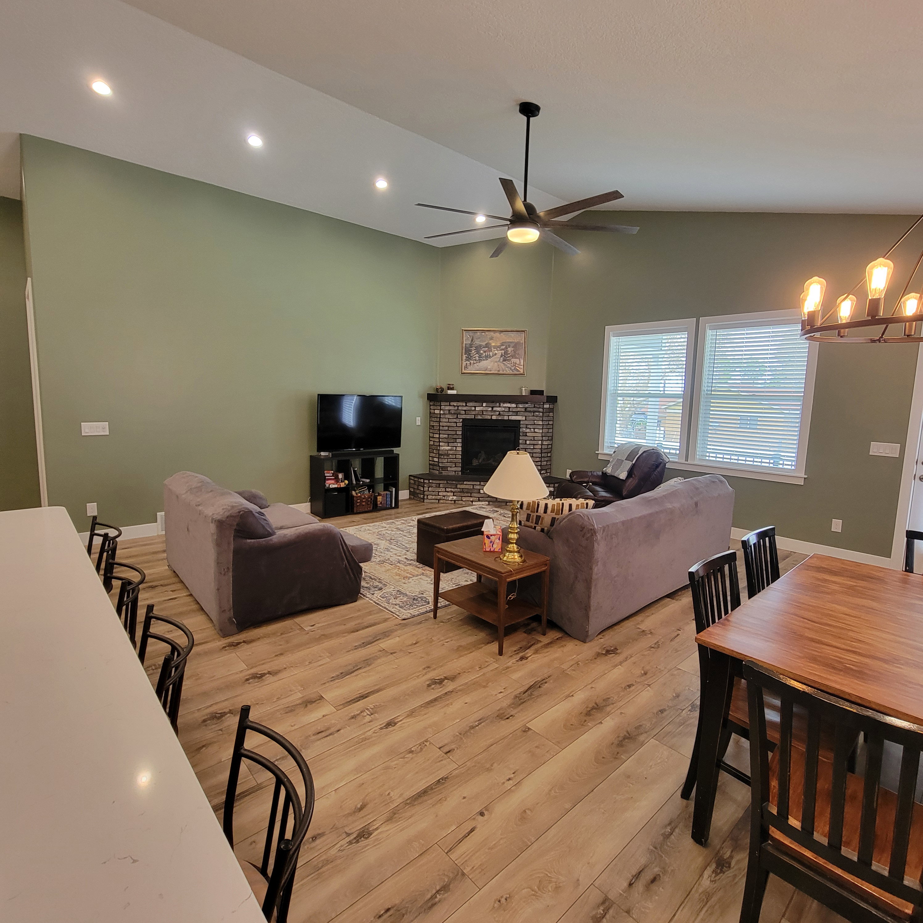 Open-concept living space featuring vaulted ceilings with recessed lighting, wood-finish flooring, a brick-surround fireplace with a wood mantle, and multiple windows with blinds
