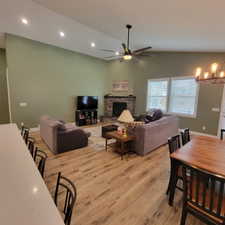 Open-concept living space featuring vaulted ceilings with recessed lighting, wood-finish flooring, a brick-surround fireplace with a wood mantle, and multiple windows with blinds