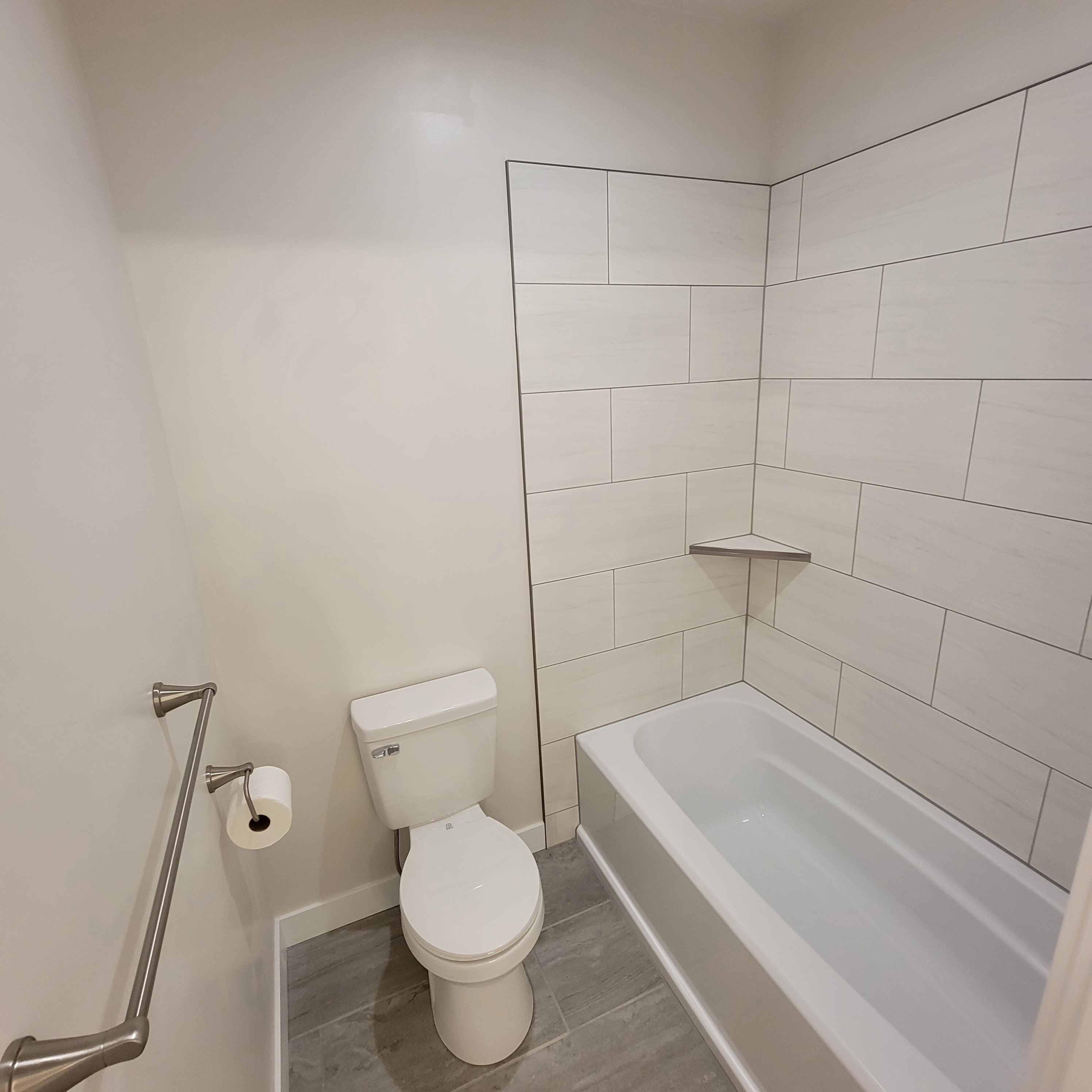Contemporary bathroom featuring a white bathtub, light-toned wall tiling, a built-in shower shelf, and wood-finish flooring