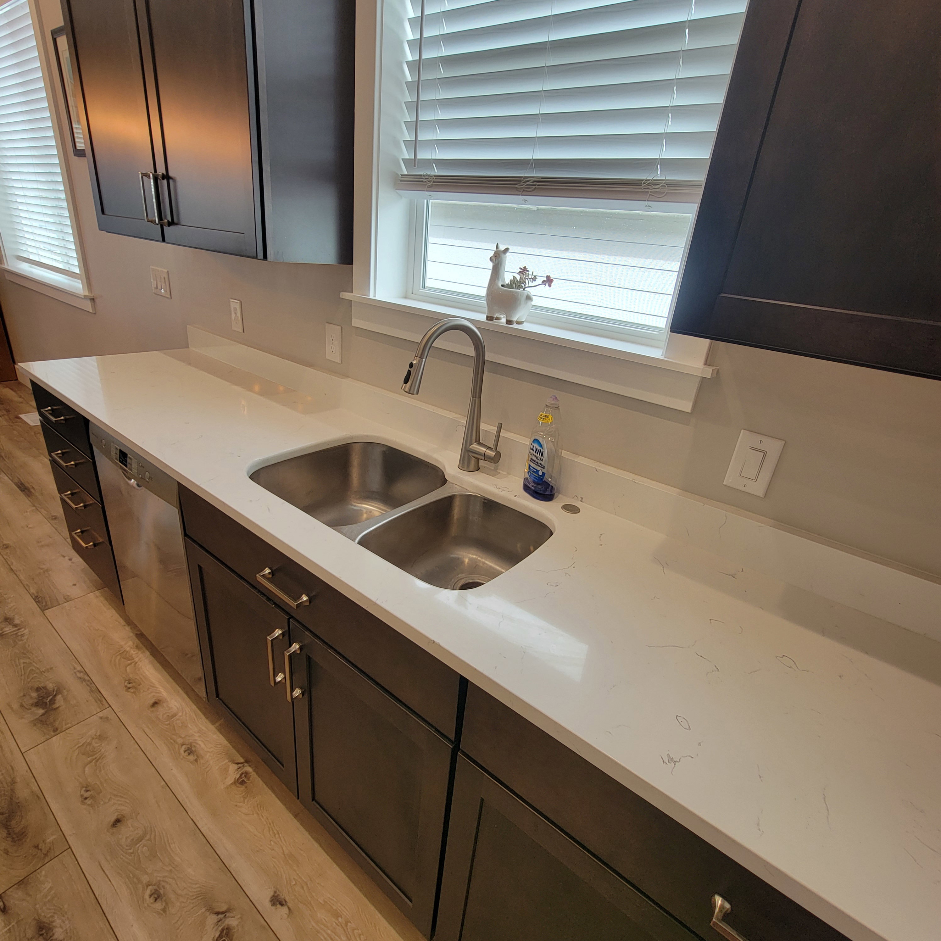 Kitchen featuring wood-finish flooring, dark cabinetry with brushed nickel hardware, light-toned countertops, stainless steel double-basin sink, and a modern pull-down faucet