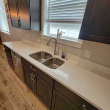 Kitchen featuring wood-finish flooring, dark cabinetry with brushed nickel hardware, light-toned countertops, stainless steel double-basin sink, and a modern pull-down faucet