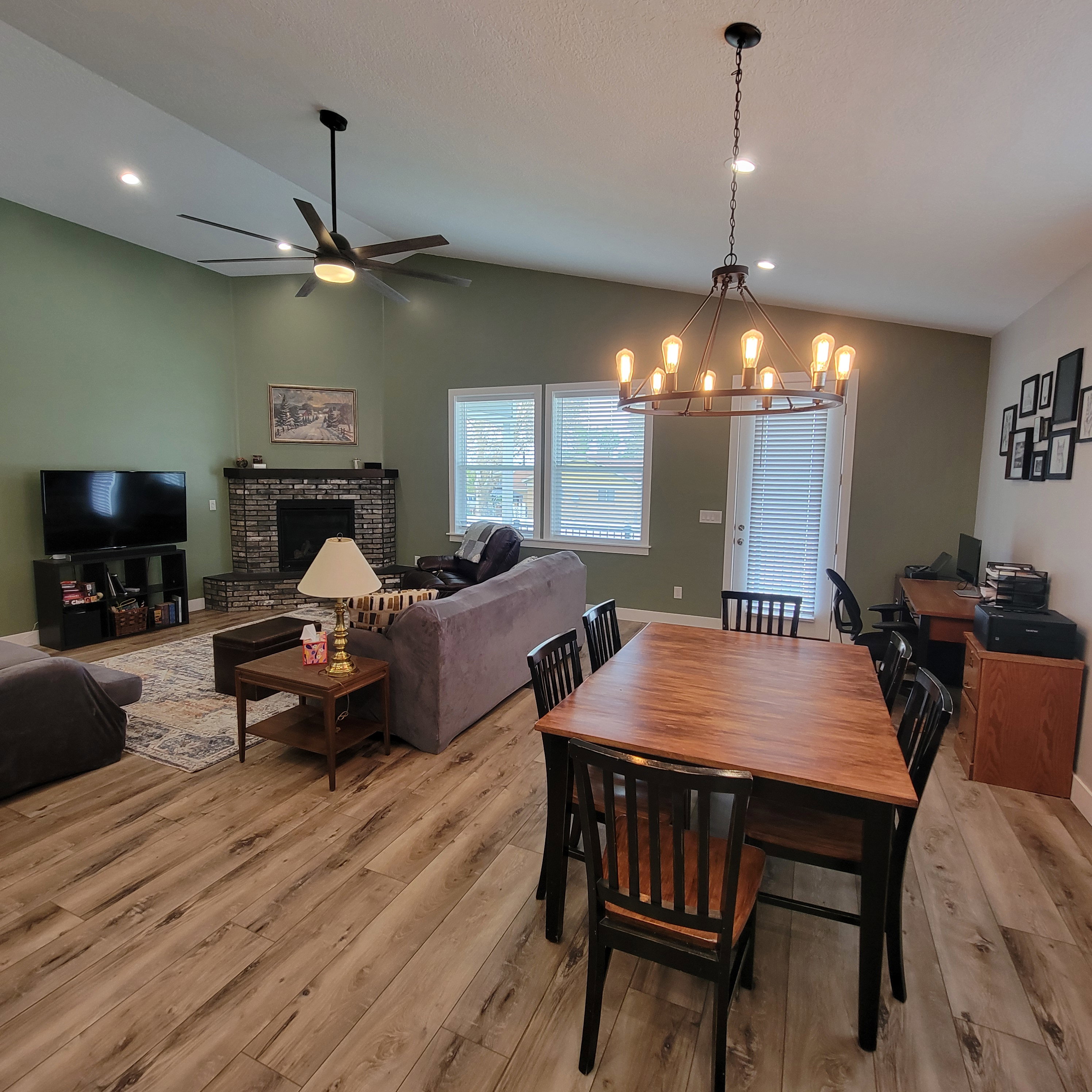 Open-concept living and dining area featuring vaulted ceilings, wood-finish flooring, a brick-surround fireplace, and recessed lighting