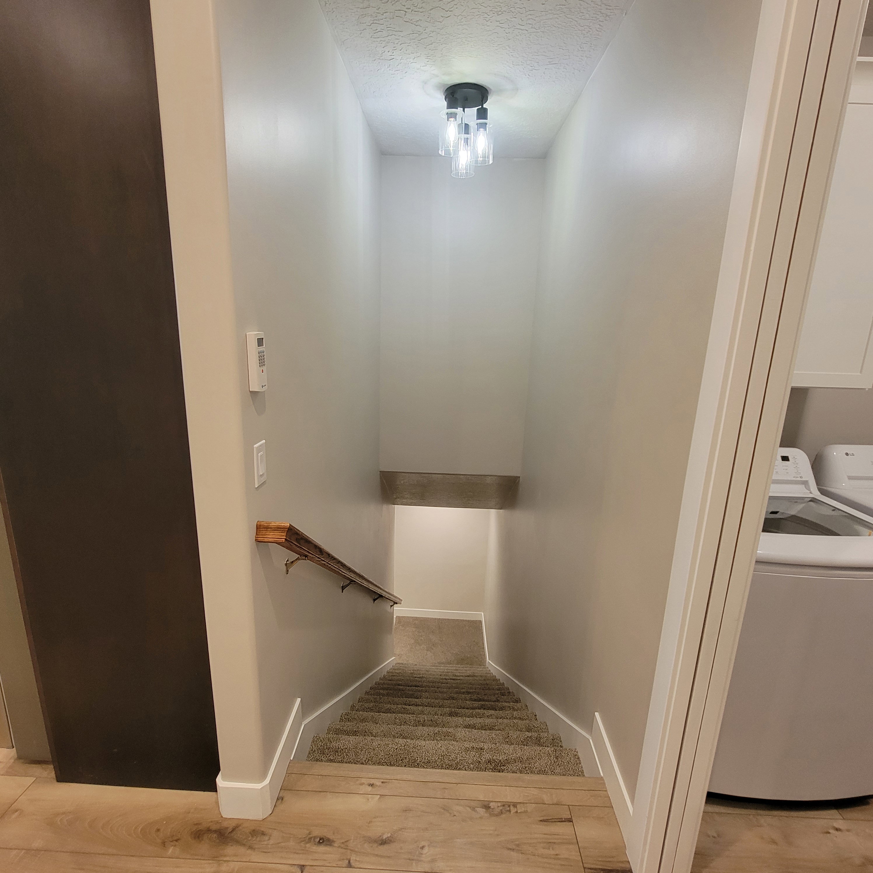Carpeted staircase descending from a wood-finish flooring landing, featuring a contemporary ceiling light fixture, a wooden handrail, and white baseboards