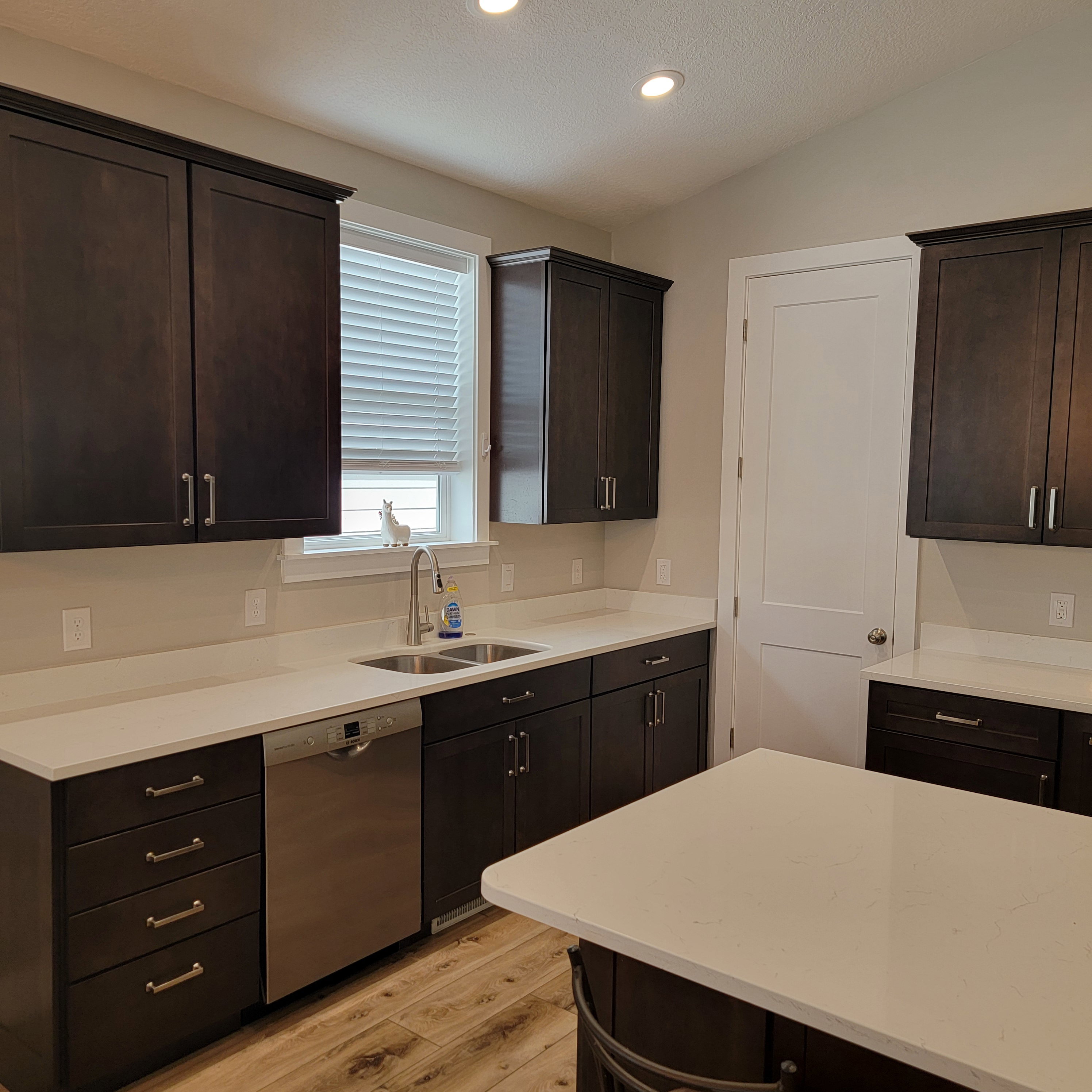 Kitchen featuring dark wood cabinetry, light countertops, a double basin stainless steel sink, a stainless steel dishwasher, and wood-finish flooring