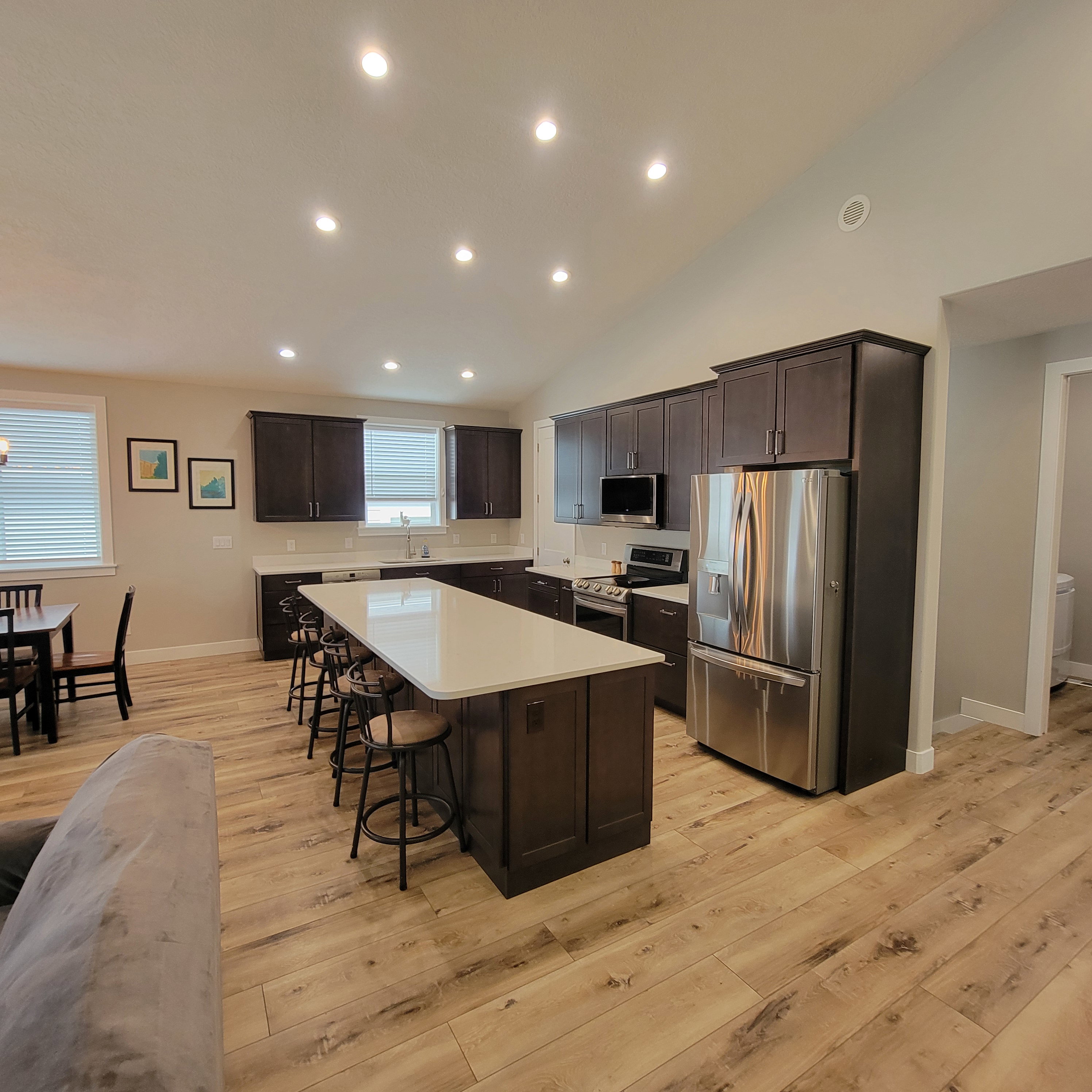 Open-concept kitchen featuring wood-finish flooring, dark wood cabinetry, stainless steel appliances, a large central island with a light-colored countertop, and recessed lighting