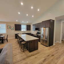 Open-concept kitchen featuring wood-finish flooring, dark wood cabinetry, stainless steel appliances, a large central island with a light-colored countertop, and recessed lighting